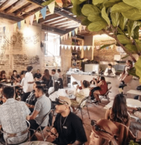 A group of people sit at tables in a decorated café in Bogotá, featuring exposed brick walls, bunting, and a visible kitchen area.